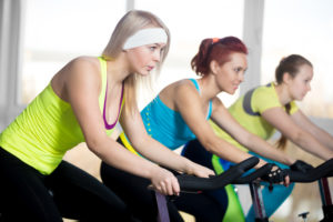 Fitness practice, group of three fit beautiful females cycling in sports club, doing cardio exercises for slimming in class, focus on blond woman controlling her breath